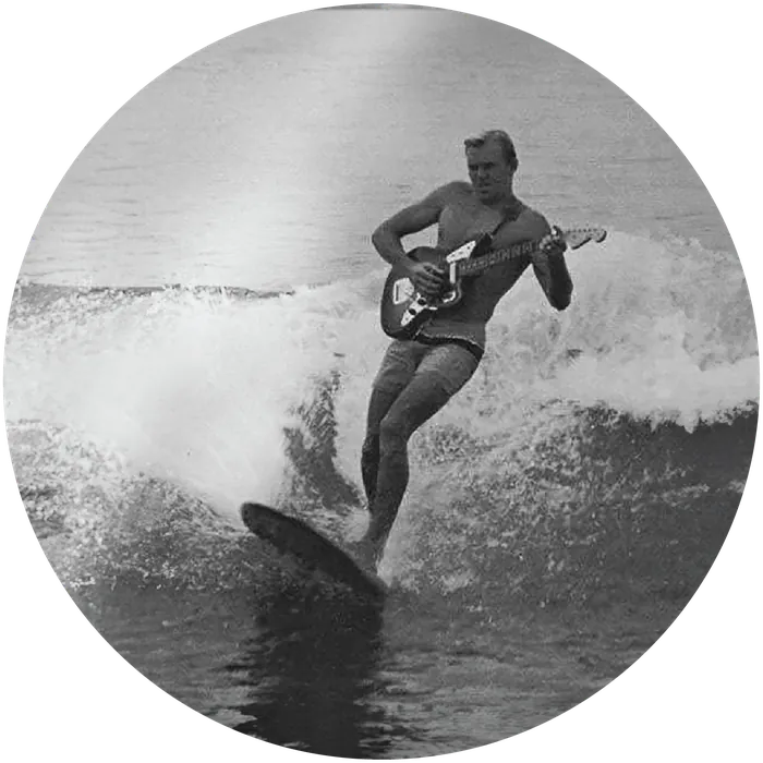 Vintage black-and-white photo of a surfer playing guitar while riding a wave