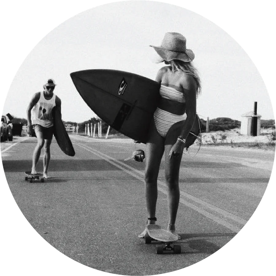 Two friends carrying surfboards and walking down a street on skateboards